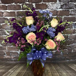 Bouquet of cream roses, purple flowers, and blue blooms in a glass vase with a ribbon