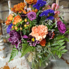 Colorful mixed bouquet with orange roses, peach carnation, and purple blooms in a clear glass vase.