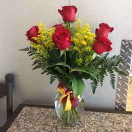 Arrangement of red roses with yellow filler flowers in a clear glass vase with a burgundy and yellow bow