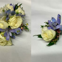 Two floral boutonnieres with cream roses and purple flowers on white fabric.
