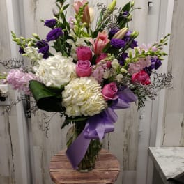 Tall mixed bouquet of pink, white, and purple flowers in a glass vase with a purple ribbon