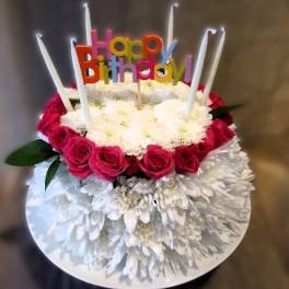 Round cake-shaped arrangement of white mums and pink roses with tall candles and a Happy Birthday pick