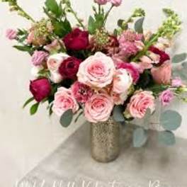 Pink and red rose arrangement with accent blooms in a tall metallic vase on a light surface.