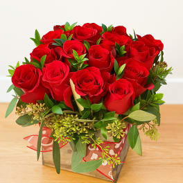 Red rose arrangement in a square glass vase with greenery and ribbon