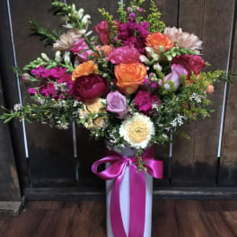 Mixed bouquet of pink, orange, and white flowers in a tall white vase with a pink ribbon
