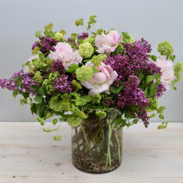 Loose garden-style bouquet of pink peonies, purple lilacs, and green blooms in a clear glass cylinder vase