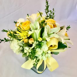 Short arrangement of yellow roses, white lilies, and green hydrangeas in a square glass vase with a yellow ribbon bow