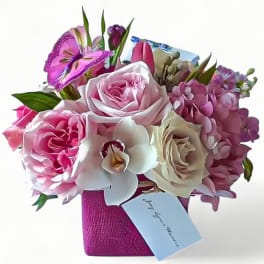 Compact pink and cream rose arrangement with orchids in a magenta cube vase and card.