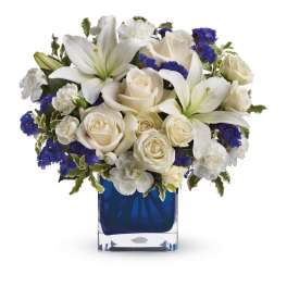 White lilies and roses arranged in a blue glass vase with blue filler flowers
