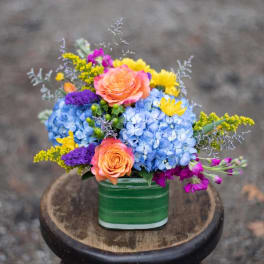 Colorful bouquet in a green square vase with blue hydrangeas and orange roses
