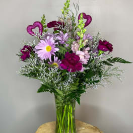 Tall vase arrangement of purple carnations, lavender daisies, and pink flowers with glitter heart accents.