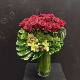 Red roses arranged in a tall glass vase with green orchids and tropical leaves