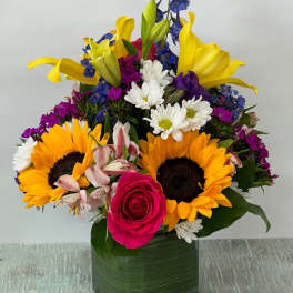 Colorful mixed bouquet in a square glass vase with sunflowers, lilies, roses, and daisies