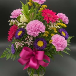 Mixed bouquet of pink carnations, gerbera daisies, and purple asters in a glass vase with a pink bow