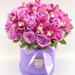 Pink roses and orchids arranged in a lavender hat box with a ribbon