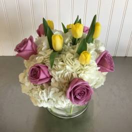 Compact arrangement of yellow tulips, lavender roses, and white hydrangeas in a clear glass vase