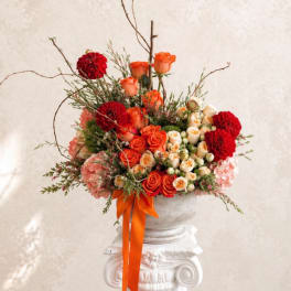 Orange and red rose arrangement in a white urn with an orange ribbon