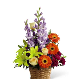 Tall mixed arrangement with orange gerbera daisies, yellow roses, green lilies, and purple flowers in a basket