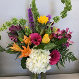 Tall mixed bouquet with white hydrangea, orange lilies, pink gerbera, yellow blooms and green Bells of Ireland in a clear vase
