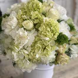 Round arrangement of white and pale green hydrangeas, roses, and carnations in a white vase