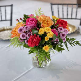 Bright mixed arrangement of roses, carnations, and daisies in a clear glass vase on a table.
