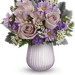 Lavender roses and purple daisies with white filler flowers in a ribbed pale purple glass vase