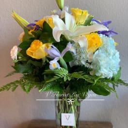 Tall arrangement of white lilies, yellow roses, blue hydrangeas, and purple iris in a clear glass vase
