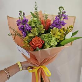 Handheld bouquet of purple, orange, yellow, and green flowers wrapped in peach paper