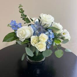 Low arrangement of white roses and blue flowers in a clear glass vase with a small starfish accent.