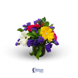 Short bouquet of yellow, red, pink, and white gerbera daisies with purple filler in a glass vase