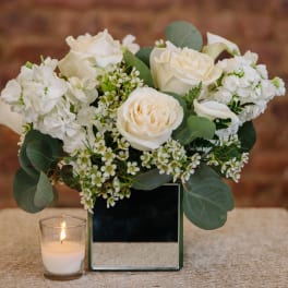 White rose arrangement in a mirrored square vase with a small candle beside it