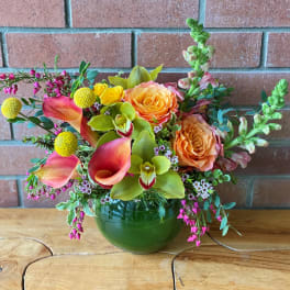 Low arrangement of orange roses, pink calla lilies, and green orchids in a round green glass vase