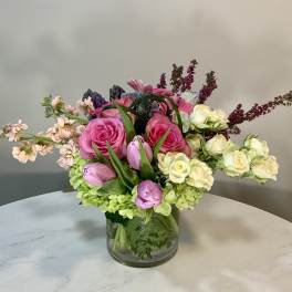Low glass vase of pink roses, purple tulips, cream spray roses and mixed blooms.