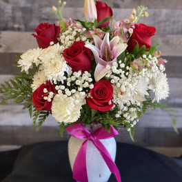 Bouquet of red roses, pink lilies, and white daisies in a white vase with a pink ribbon