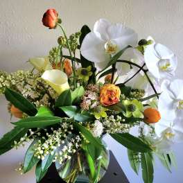 Arrangement of white orchids, yellow calla lilies, and orange ranunculus in a clear glass vase