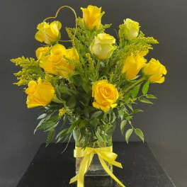 Tall arrangement of yellow roses in a clear glass vase with a yellow bow and heart-shaped accent