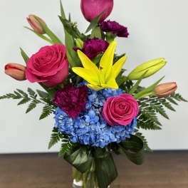 Bright bouquet with pink roses, yellow lilies, blue hydrangea, and purple carnations in a glass vase