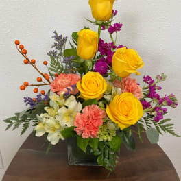Mixed bouquet of yellow roses, pink carnations, and purple flowers in a glass vase