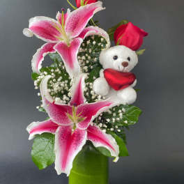 Pink lilies and red roses in a green vase with a teddy bear holding a heart