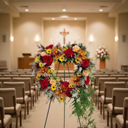 Standing round wreath of red roses, yellow daisies and striped mums on an easel in a chapel aisle