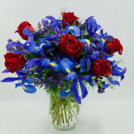 Arrangement of red roses and blue irises with purple filler flowers in a clear glass vase