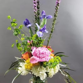 Tall mixed flower arrangement in a glass vase with pink, purple, white, and orange blooms