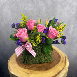 Pink roses and purple filler flowers arranged in a moss-covered box with a striped ribbon.