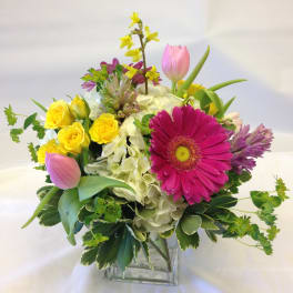 Mixed bouquet in a square glass vase with pink tulips, yellow roses, and a magenta gerbera daisy