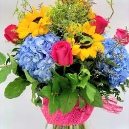 Tall mixed bouquet with yellow sunflowers, pink roses, and blue hydrangeas in a clear glass vase