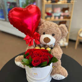 Teddy bear holding a red heart balloon beside red roses in a white box