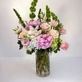 Tall pastel arrangement of pink peonies and roses with green Bells of Ireland in a clear glass vase