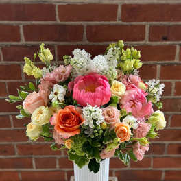 Tall arrangement of pink peonies, peach and orange roses, and white blooms in a white ribbed vase