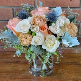 Peach and cream roses with blue thistle-style blooms arranged in a clear glass vase