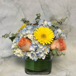 Bouquet of blue and white hydrangeas with yellow gerbera daisies and peach roses in a glass vase